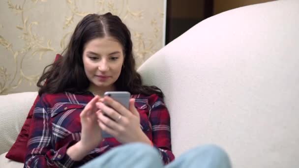 Jolie femme riante utilisant le téléphone mobile, se détendre sur le canapé, maison confortable — Video