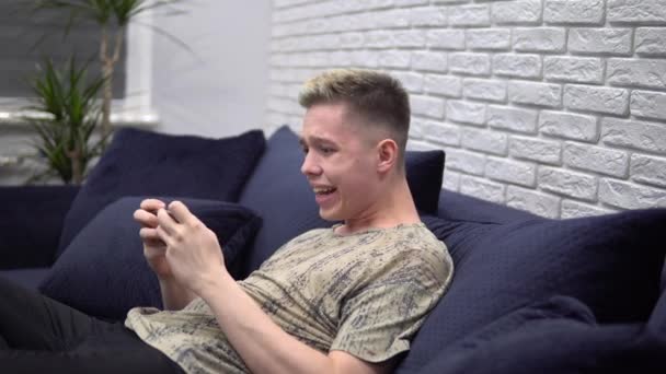 Привлекательный счастливый человек играет в мобильные игры на смартфоне, расслабляется, отдыхает дома — стоковое видео
