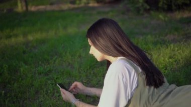 Parkta çimlerde dinlenen çekici bir kadın akıllı telefon kullanarak yazın tadını çıkarıyor.