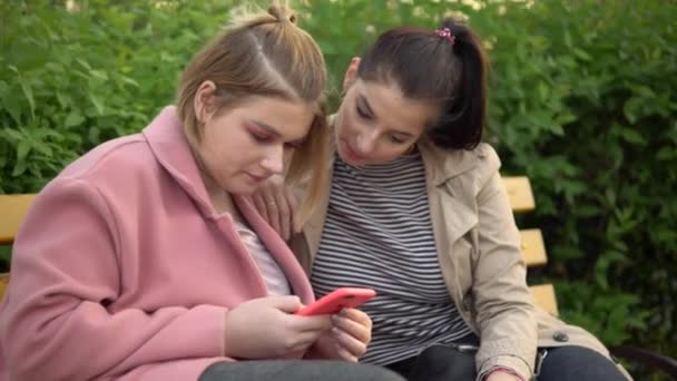 Дві молоді щасливі дівчата дивляться на мобільний екран, друзі сидять на лавці в парку — стокове відео