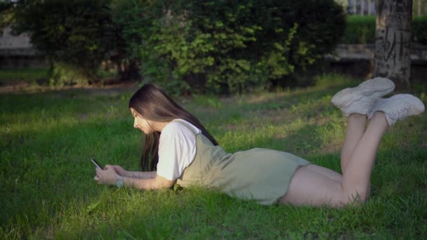 Mulher relaxante encontra-se na grama no parque e usando smartphone — Vídeo de Stock