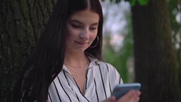 Жінка використовує мобільний телефон, відпочиває в парку, сидить під деревом — стокове відео