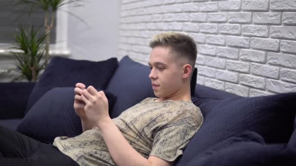 Jovem jogando jogo móvel, usando telefone, relaxando no sofá em casa — Vídeo de Stock