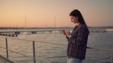 Gün batımında su kenarında akıllı telefon sohbeti yapan çekici bir kadın.