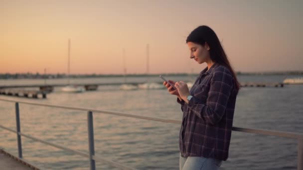 Vonzó nő használ okostelefon chat szabadban, naplementekor, közel a vízhez — Stock videók