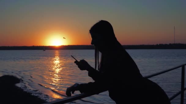 Femme à l'aide d'un smartphone, promenade en soirée sur la jetée, silhouette de la femme au coucher du soleil — Video