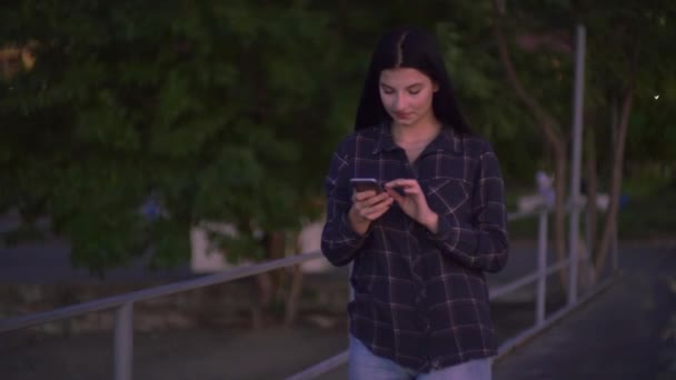 Sokakta yürüyen ve akıllı telefon kullanan bir kadın. Ekrana, gece şehrine bakıyor. — Stok video