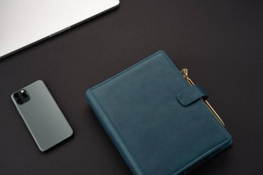 Yeşil el yapımı defterleri, gri dizüstü bilgisayarı, akıllı telefonu ve altın kalemi olan siyah masa.