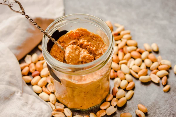 Peanut paste in an open jar and and spoon, peanuts on the gray t