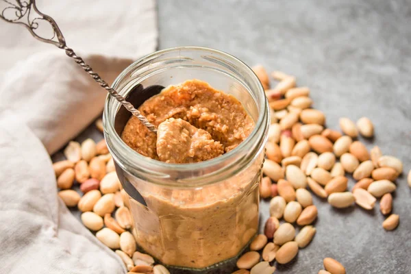 Peanut paste in an open jar and and spoon, peanuts on the gray t