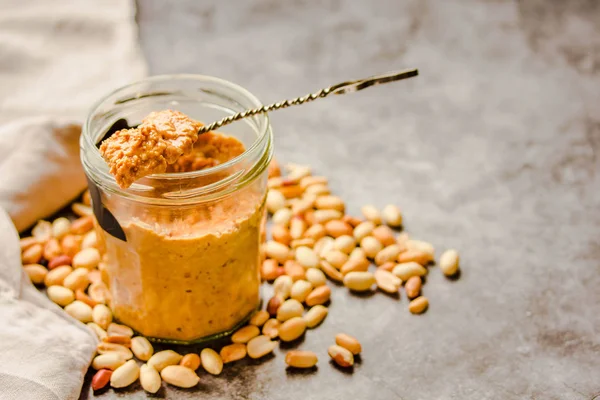 Erdnusspaste in einem offenen Glas und einem Löffel, Erdnüsse auf dem grauen T — Stockfoto