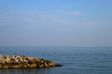 Limanın girişindeki taştan dalgalanma suyu. Taş iskele ve mavi deniz seviyesi. Mavi yüzeyde küçük dalgalanmalar. Açık beyaz bulutlu okyanus ve gökyüzü ufku. Rahatlama ve seyahat.