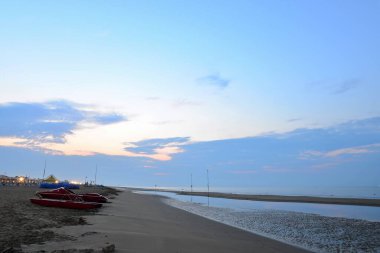 Bir İtalyan sahilinde şafak vakti. Kumsalda kırmızı cankurtaran botları. Dalgalar alçalınca deniz yatağı açıkta kalıyor. Yükselen güneş, bulutlu mavi gökyüzü. Turistler olmadan temiz sahil. Rahatlamaya hazır..