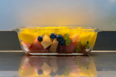 Taze meyve salatalı cam kase, mutfak çalışma alanı.