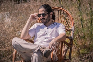 Siyah güneş gözlüklü genç bir adam hasır bir sandalyede oturur. Karizmatik bir şekilde, açık havada ve güneşli bir yaz gününde..