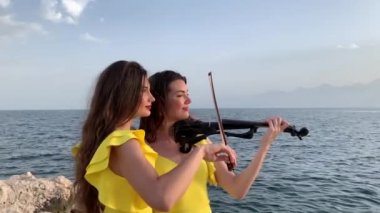 Türkiye 'nin Antalya kentinde fotoğrafçıya elektrikli kemanla poz veren sarı elbiseli iki genç ikiz kardeş kemancıya yakın çekim. Deniz, dağlar ve arka planda bulutlu gökyüzü. 4k stok video.