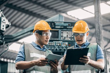 30 'lu yaşlarda iki Asyalı işçi metal fabrikasında konuşuyorlar. Kasketli ve koruyucu gözlüklü. Bilgisayarı gösteren ve modern fabrikada yeni planlardan bahseden adam..