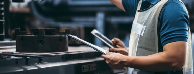 Elinde tablet ve belge planıyla bir fabrikada çalışan iki işçi fabrikadaki endüstriyel makinelerde çalışmak isteyen bir mühendis..