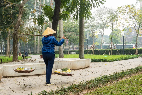 An elderly Vietnamese woman wearing a traditional cone hat walks with a traditional don ganh wicker basket yoke in a park. Street food vendor in Hanoi