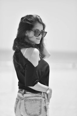 Siyah tişörtlü, moda güneş gözlüklü ve kot pantolonlu seksi genç kadın sahilde denize bakıyor. Sahilde güneşlenen bir kız..