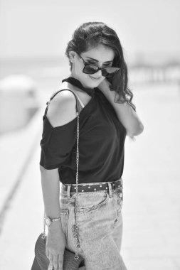 Siyah tişörtlü, moda gözlüklü, kırmızı çantalı, kot pantolonlu seksi genç kadın, sahilde dur. Sahilde güneşlenen bir kız..