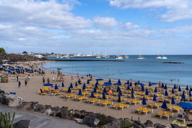 Lanzarote 'de canlı bir plaj sahnesi. Mavi bir gökyüzünün altında, güneş yatakları, şemsiyeler ve yelkenliler var. Lanzarote, İspanya, 2025-07-20