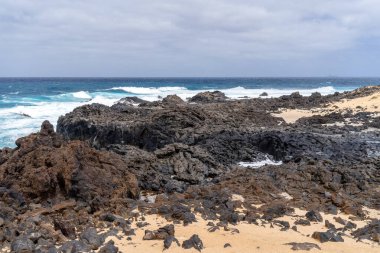 Lanzarote, La Graciosa adasındaki manzarada volkanik kayalar, kumlu kumsallar ve bulutlu bir gökyüzü altında okyanus dalgaları var..