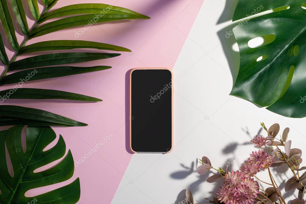 Flat lay of a pink smartphone, palm and monstera leaves, and pink flowers on white and pink background