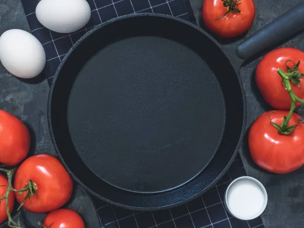 Black cast-iron frying pan on black cloth, white chicken eggs, red tomatoes and salt. Black background with blank space for text. Top view. Copy space.
