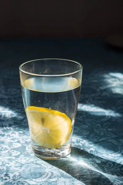 Färskt rent vatten i ett glas med skivor av citron. Lemonad på en vacker blå duk i ljusa morgonsolen. Levande vatten, hälsosam dryck — Stockfoto