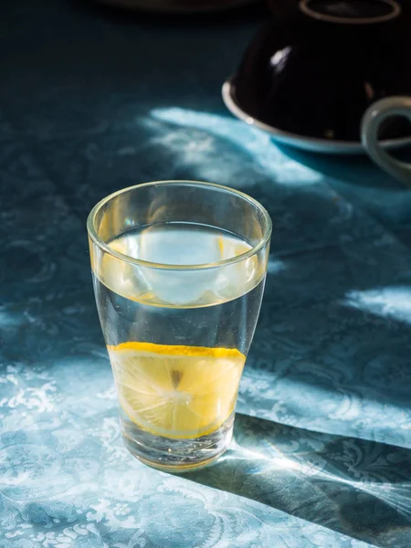 Свежая чистая вода в стакане с ломтиками лимона. Лимонад на красивой голубой скатерти под ярким утренним солнцем. Живая вода, здоровый напиток — стоковое фото