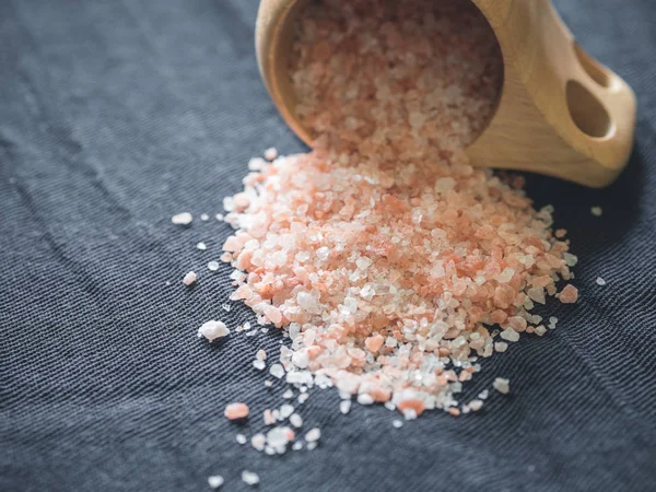 Useful spices concept. Large dark pink himalayan salt spilled from a wooden cup onto a black tablecloth. Closeup