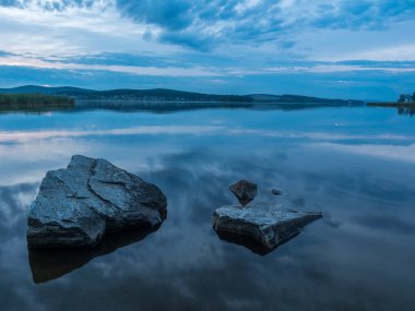 Sakin, dinginlik, meditasyon konsepti. Gölde gün batımı, ön plandaki sudaki taşlar, sessiz su, bulutsuz gökyüzü. Mavi saat