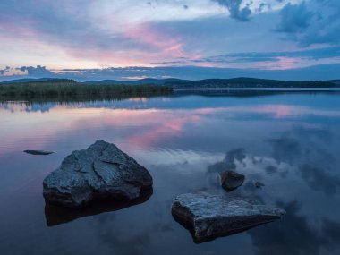Sakin, dinginlik, meditasyon konsepti. Gölde gün batımı, ön plandaki sudaki taşlar, sessiz su, bulutsuz gökyüzü. Mavi saat