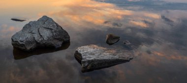 Sakin, dinginlik, meditasyon konsepti. Gölde gün batımı, ön plandaki sudaki taşlar, sessiz su, bulutsuz gökyüzü. Altın saat