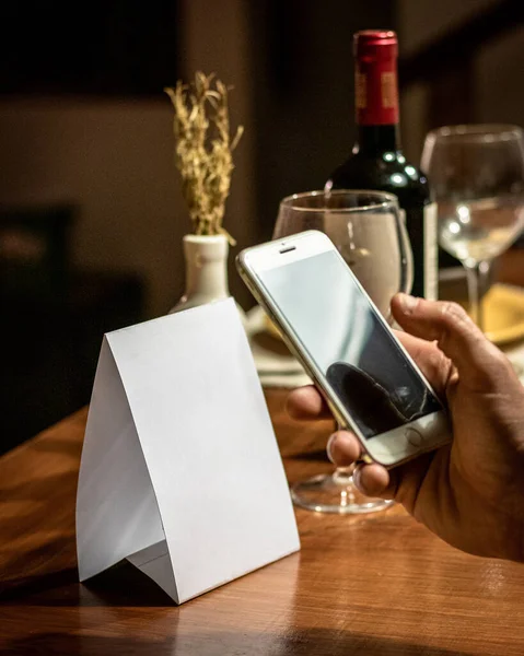Cep telefonu. Restoran masasındaki boş QR kartını tarıyor. Arka planda gözlük ve şarap şişesi var.