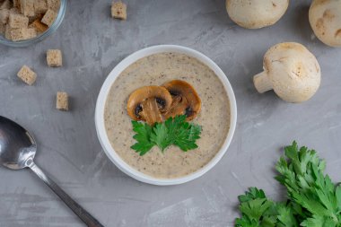 Beyaz kasedeki kremalı mantar çorbası mantar dilimleri ve yeşil maydanozla süslenmiş taze çiğ mantarlar, maydanoz, kurutulmuş ekmek ve beton yüzeyde kaşık