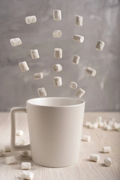 White plain simple cup standing on light wooden table surrounded by flying white marshmallows at kitchen