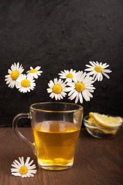 Şeffaf cam bir fincanda limonlu yeşil çay koyu kahverengi ahşap bir masada duruyor. Etrafı uçan beyaz papatya çiçekleriyle çevrili. Dikey yönelim