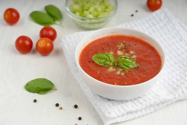 Red gazpacho soup with green basil leaves in a bowl standing on napkin on white wooden table surrounded by peppercorns, tomatoes and chopped cucumber. Horizontal orientation