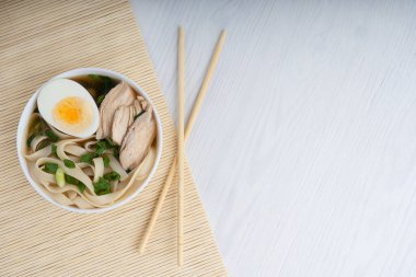 Erişteli tavuk suyundan yapılmış Asya ramen çorbası. Yarım yumurta ve hasırın üzerindeki et dilimleri ile beyaz ahşap masada yemek çubukları ile süslenmiş. Kopyalama alanı olan resim