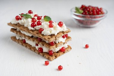 Beyaz ahşap arka plandaki kafede sunulan nane yapraklarıyla süslenmiş gevrek ekmek, krem peynir ve kırmızı frenk üzümünden yapılmış üç katlı sandviçin yakın görüntüsü. Seçici odaklı resim