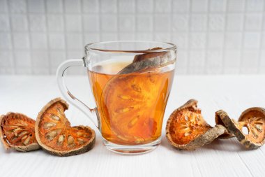 Geleneksel Thai şifalı matoom çayı kuru bael meyve dilimlerinden yapılmıştır, taş elma olarak da bilinir, şeffaf cam fincanda beyaz ahşap arka planda malzemelerle birlikte. Seçici odaklı resim