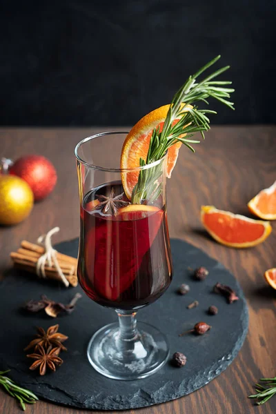 Image with selective focus of a glass of hot red mulled wine usually prepared at winter decorated with orange slices and rosemary surrounded by christmas decorations, anise, cinnamon bunch and spices