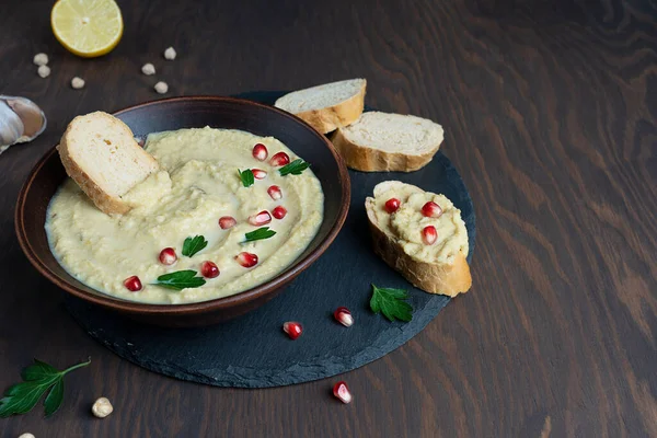 Hummus savory dish made of chickpeas with tahini, lemon juice and garlic decorated with pomegranate seeds and parsley on round tray with bread slices. on wooden background. Image with copy space