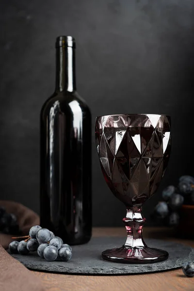 Carved retro style purple wineglass full of cabernet red wine and bottle with ripe isabella grape bunch on brown textile towel on dark wooden background at romantic evening. Vertical orientation