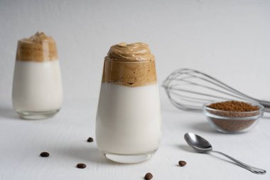 Hazır kahve tozu, şeker ve sıcak su karışımı ile yapılan ev yapımı dalgona kahvesi ile dolu bir bardak ve beyaz ahşap masa üzerinde çikolatalı şekerle servis edilen soğuk süt. Yatay