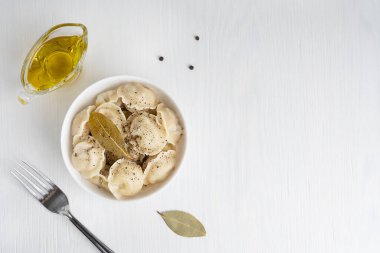 Pelmeni 'nin üst görüntüsü ya da hamur içine sarılmış kıymalı Rus hamur köfteleri beyaz ahşap zemin üzerinde siyah biberli çatal ve defne yaprağı ile servis edilir. Kopyalama alanı olan resim