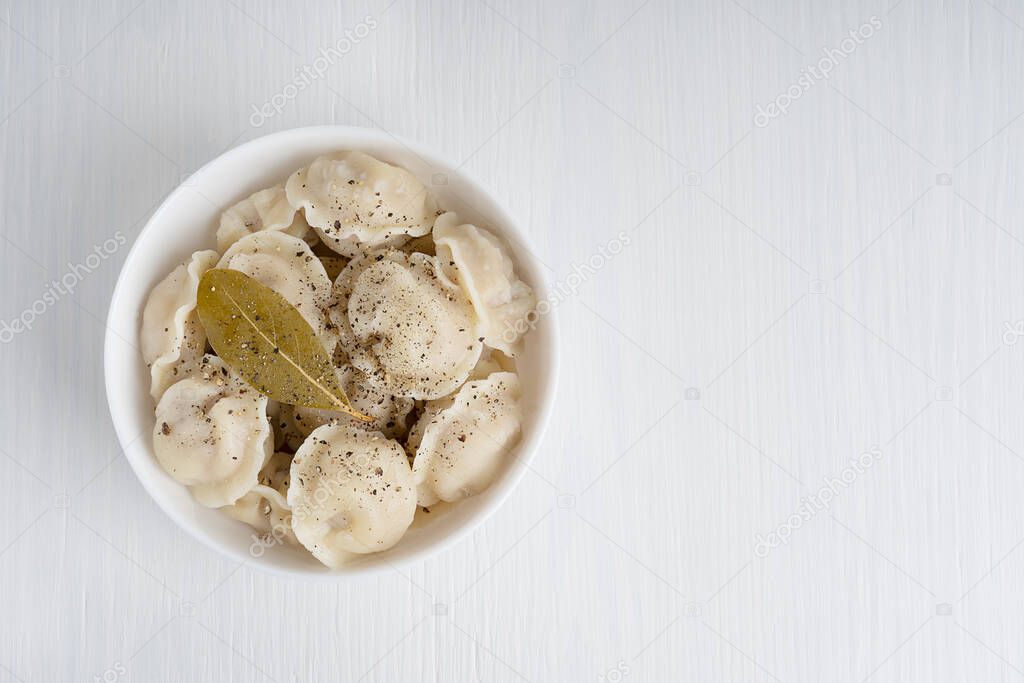 Top view of homemade Pelmeni or Russian dumplings made of minced meat filling wrapped in dough served in white bowl with black pepper and bay leaf on white wooden background. Image with copy space