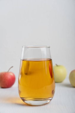 İçme bardağına dökülen şeffaf elma suyu, sağlıklı yaşam tarzı ve vejetaryen yemeklerini gösteren ham olgun meyvelerle çevrili beyaz ahşap zemin üzerinde duruyor. Dikey yönelim resmi